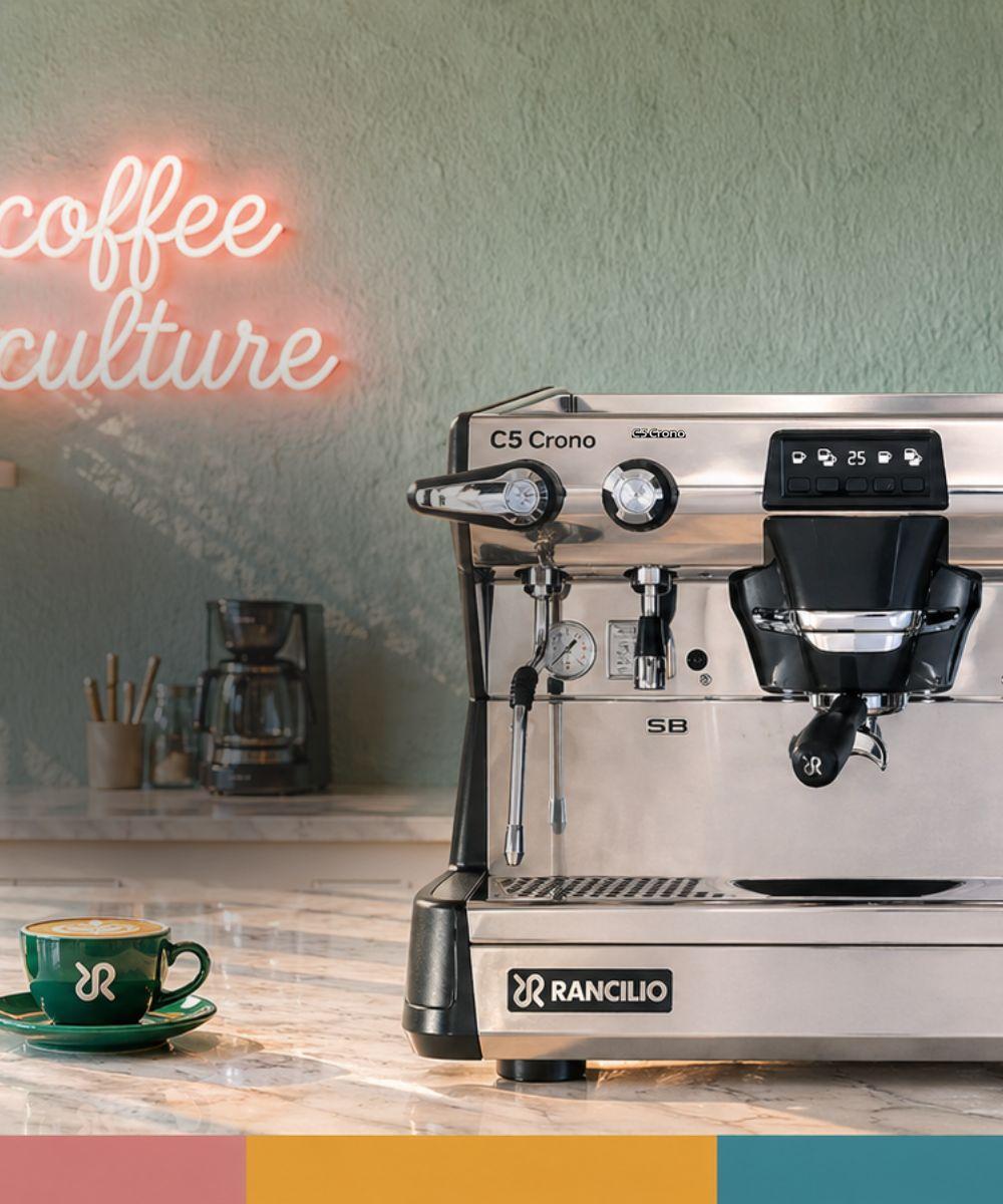 Rancilio Classe 5 CRONO espressokone kahvilaympäristössä, taustalla coffee culture valokyltti ja espressokuppi pöydällä